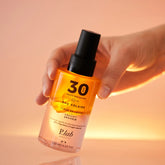 main prenant eau solaire SPF30 fond coucher de soleil rose